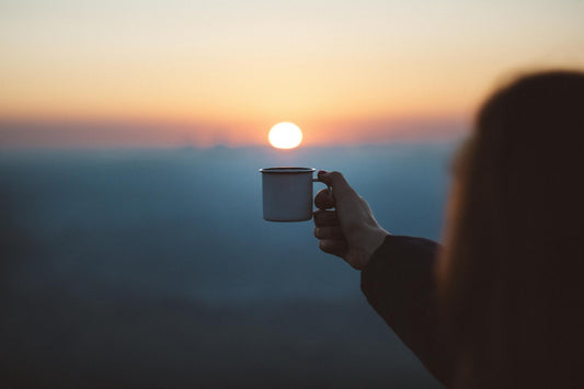 A person holding a cup of coffee up to a sunrise